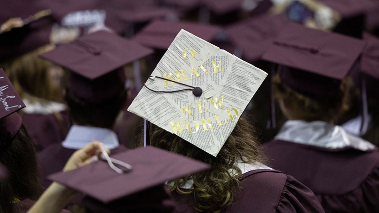 A student wears a graduation cap that is covered in book pages and says, "O Brave New World."