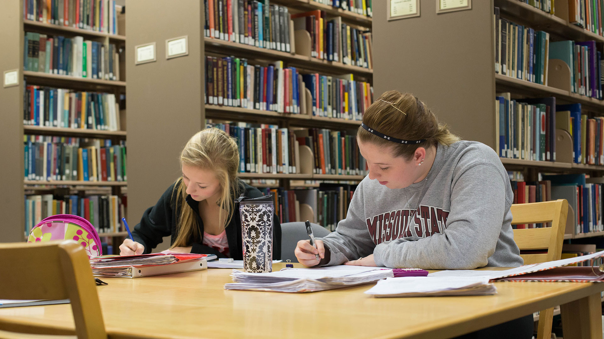 Two students sitting next to each other at a table jot down notes and complete their homework in Meyer Library.
